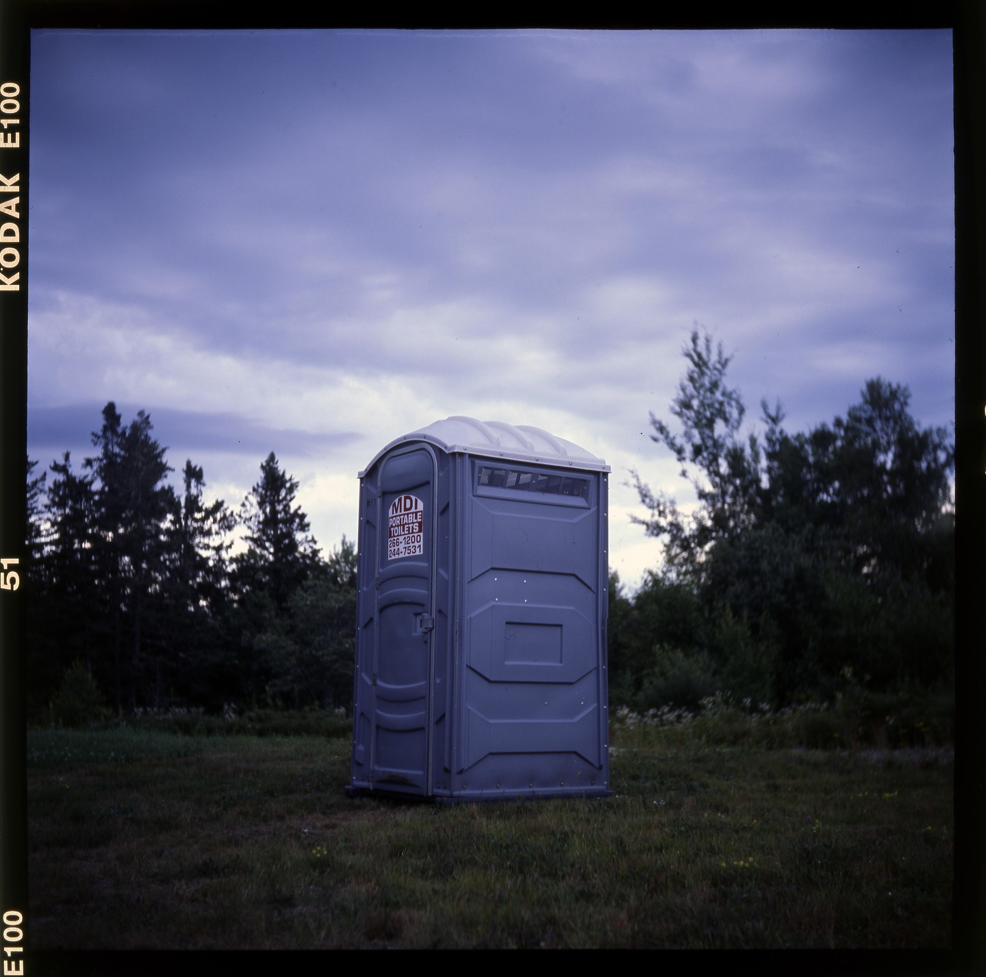 A portable toilet stands in a grassy field under a cloudy sky.