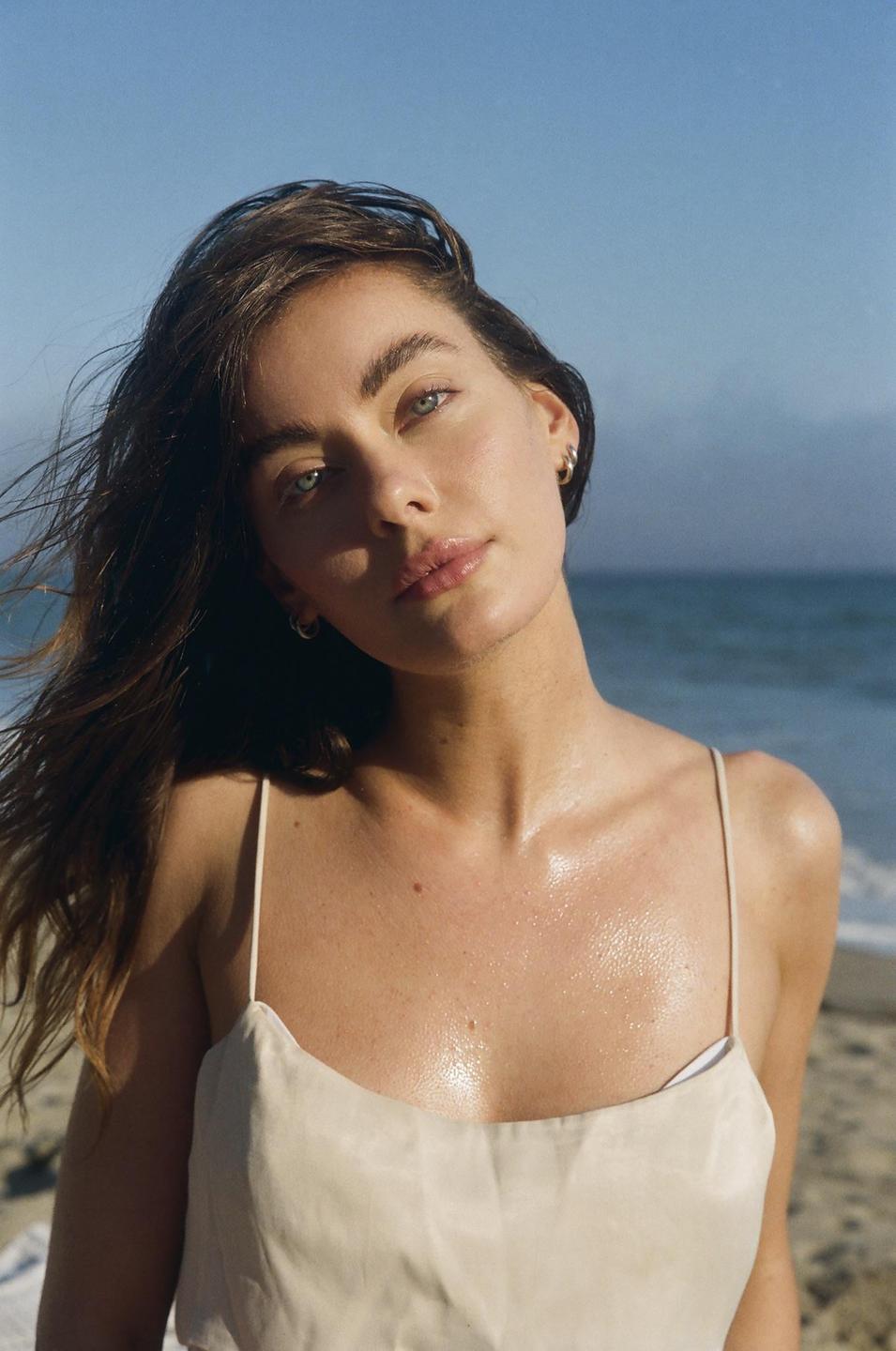 A woman with long hair and a light-colored top looks at the viewer with the ocean in the background.