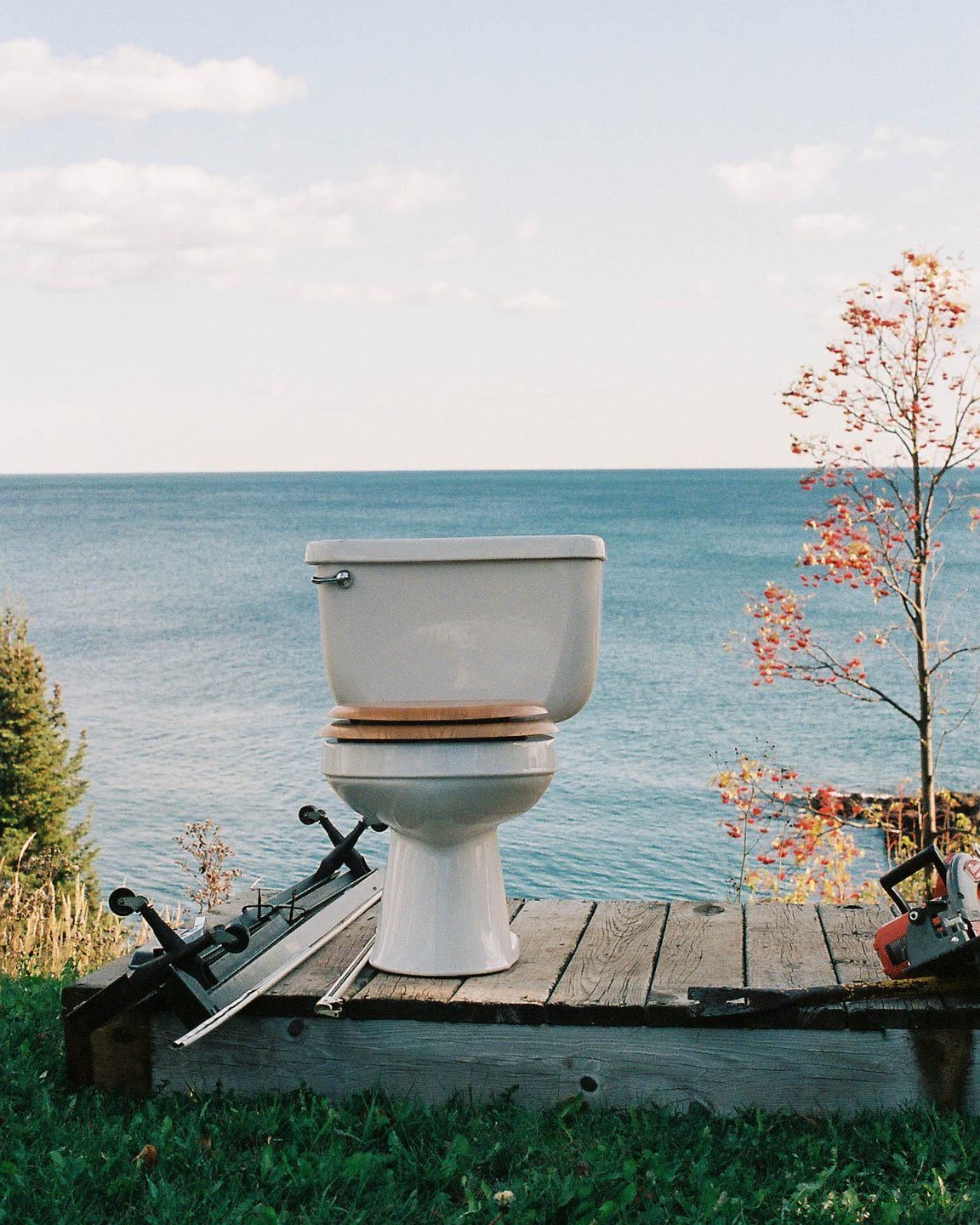 A white toilet with a wooden seat sits on a wooden platform overlooking a large body of water.