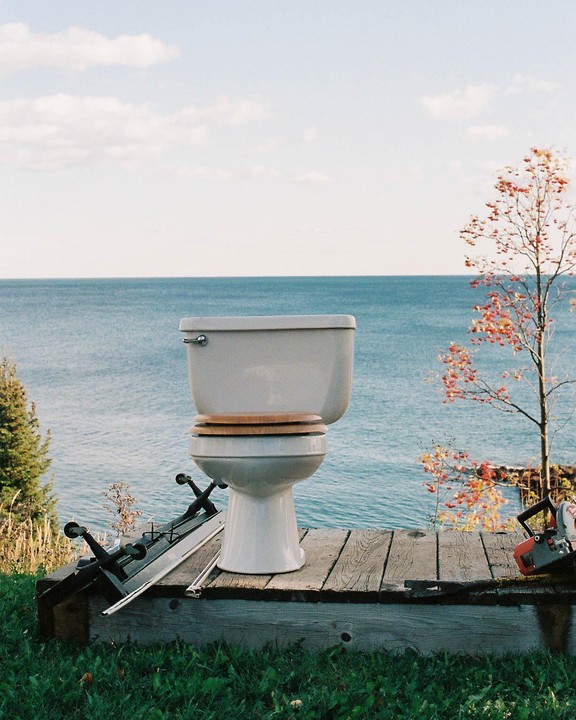 A white toilet with a wooden seat sits on a wooden platform overlooking a large body of water.
