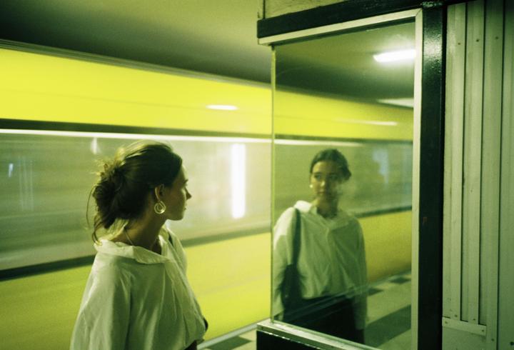 A woman with her hair tied back looks at her reflection in a glass panel.