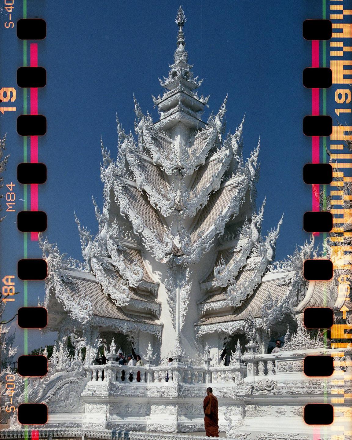 A monk stands in front of a highly ornate white temple under a clear blue sky.