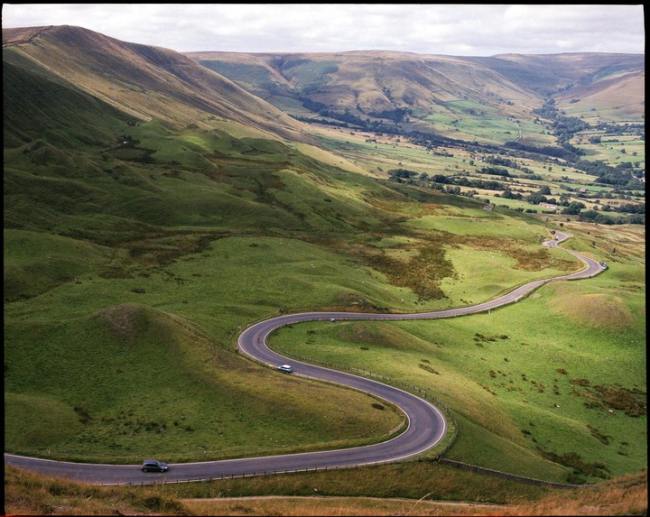 A winding road cuts through a green valley with rolling hills under a cloudy sky.