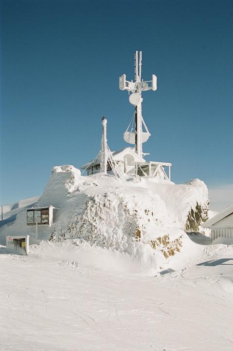 A snow-covered building with a large antenna on top sits on a snowy hill under a clear blue sky.