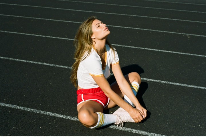 A woman sits on a running track with her head tilted back, eyes closed, and face towards the sun.