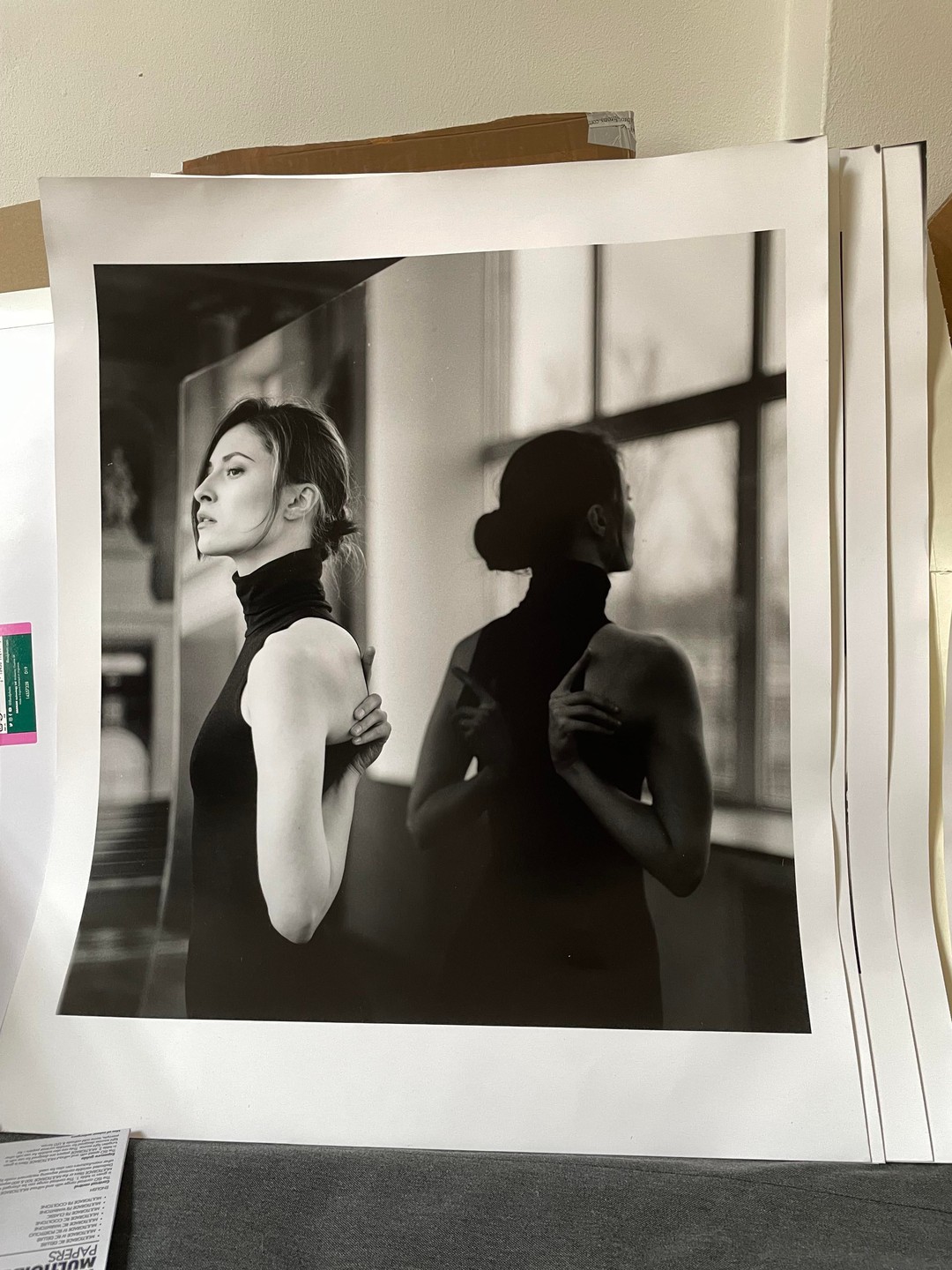 A woman in a black turtleneck stands with her back to a window, her reflection visible beside her.
