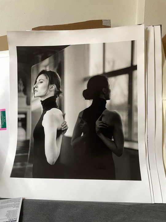 A woman in a black turtleneck stands with her back to a window, her reflection visible beside her.