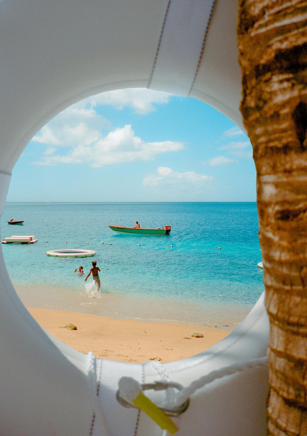 A beach scene with people in the water and a boat on the ocean is framed by a white life preserver and a palm tree.