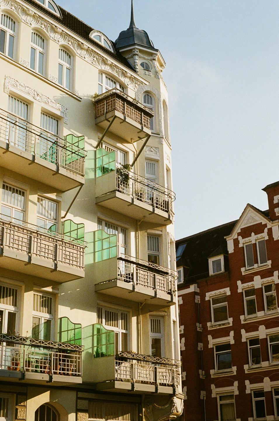 A cream-colored building with balconies and a turret stands next to a red brick building.