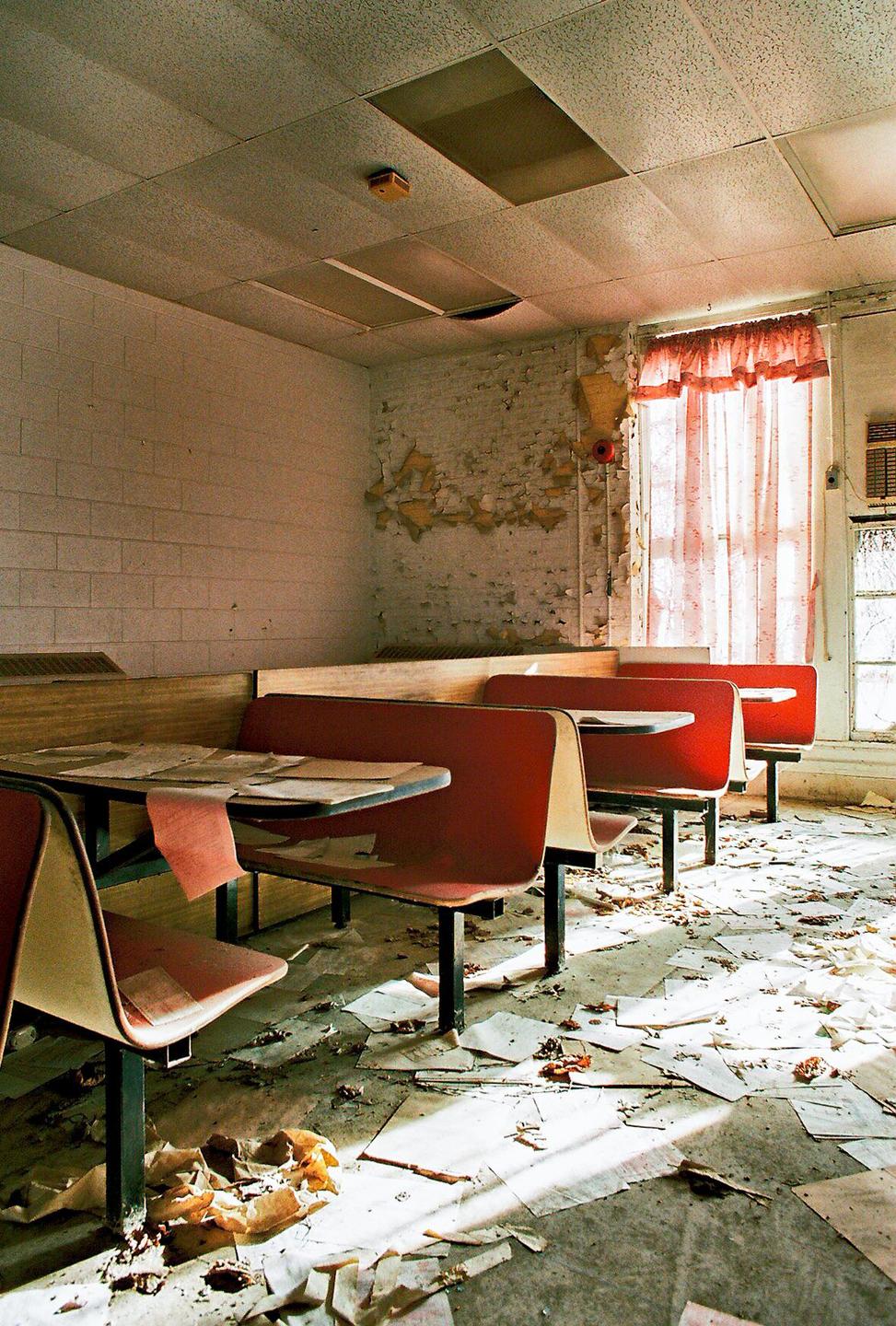An abandoned diner interior shows red booths and tables covered in debris, with peeling paint on the walls and papers scattered across the floor.