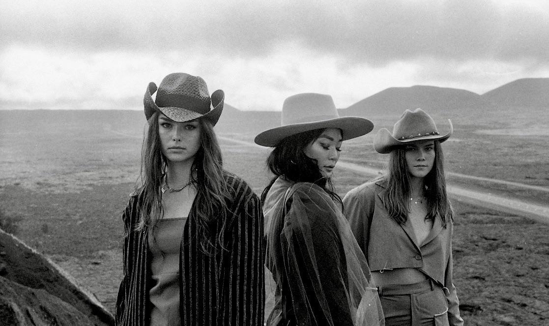 Three women wearing hats stand in a barren landscape with mountains in the distance.