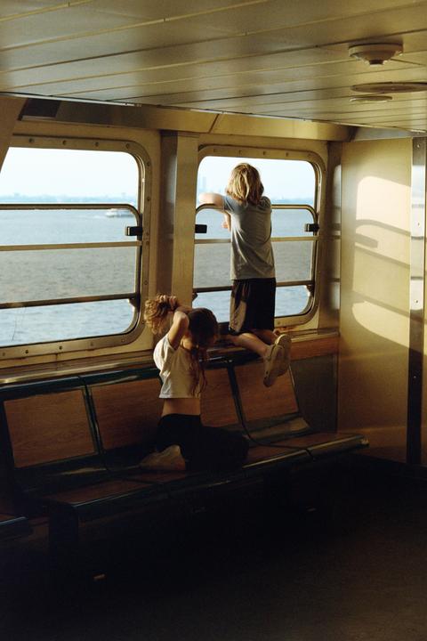 Two children are inside a boat, one looking out a window at the water and the other sitting on a bench.