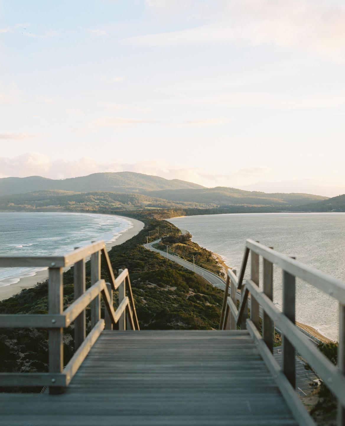 A wooden boardwalk and stairs lead down to a narrow strip of land with a road and beaches on either side, with mountains in the distance.
