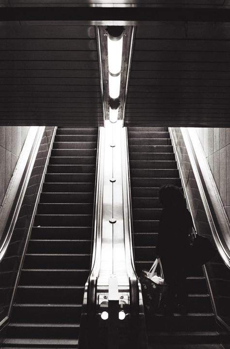 A person stands on an escalator next to another escalator going in the opposite direction, with a bright light fixture above.
