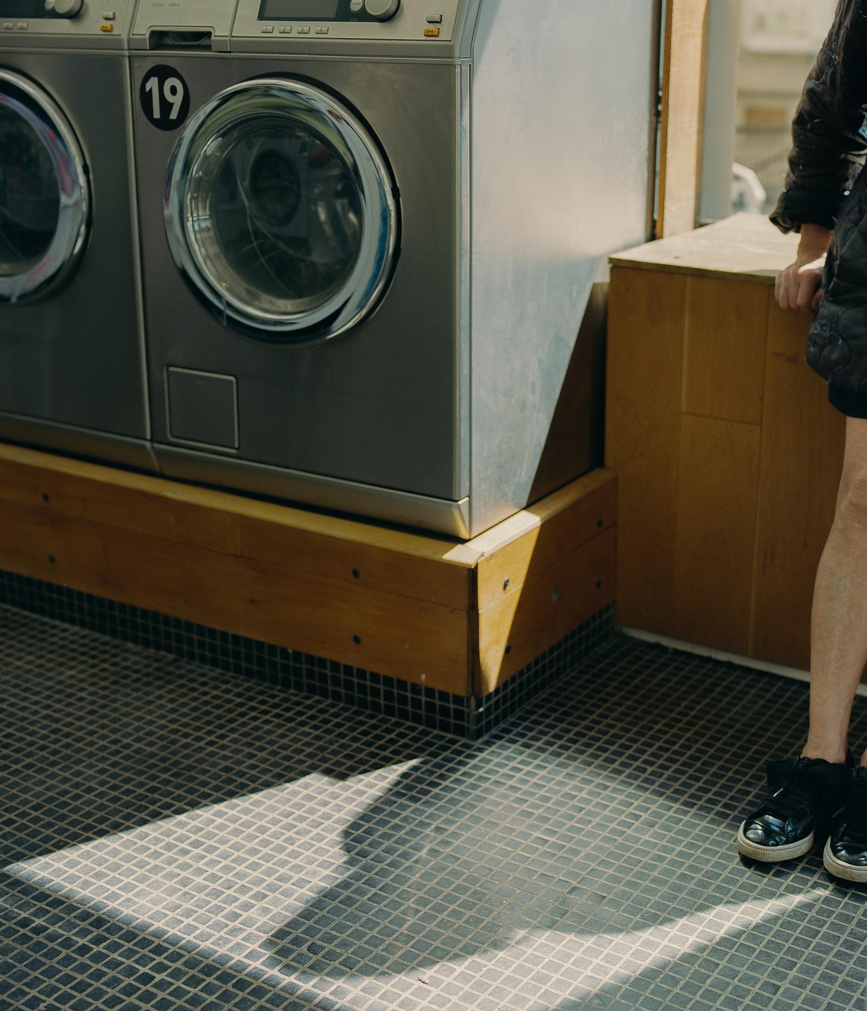 A person stands next to two washing machines in a laundromat, with sunlight casting shadows on the tiled floor.
