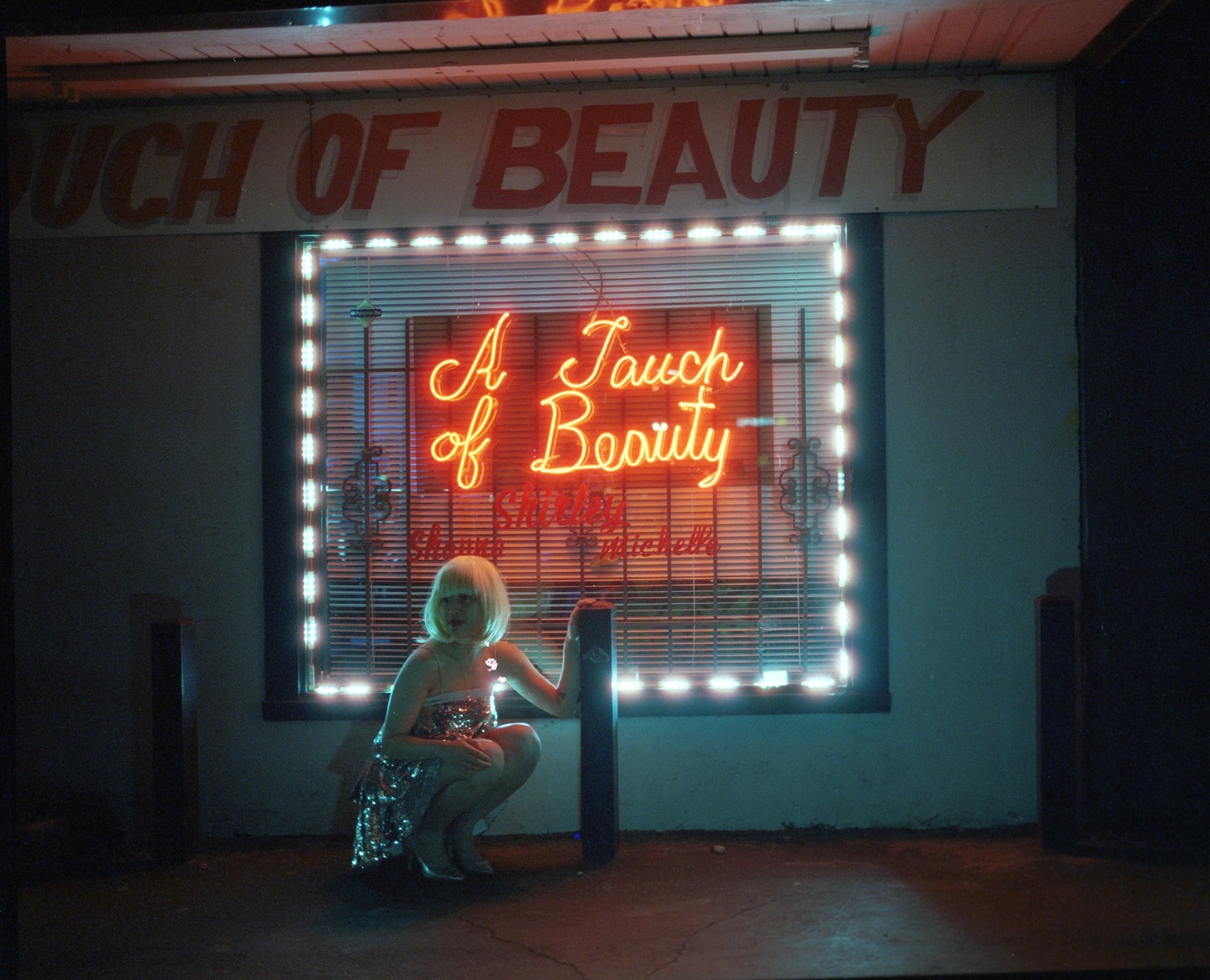 A woman in a sequin dress crouches in front of a neon sign that reads "A Touch of Beauty" at night.