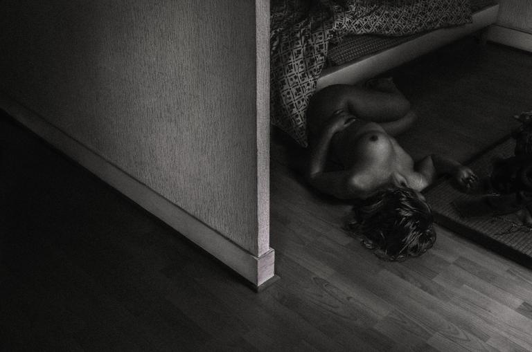 A nude woman lies on a wooden floor next to a wall.