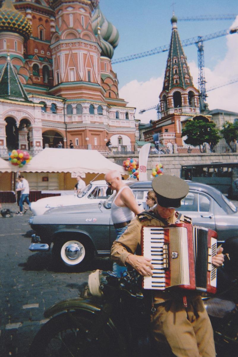 A man in a military uniform plays an accordion in front of a historic building with onion domes.