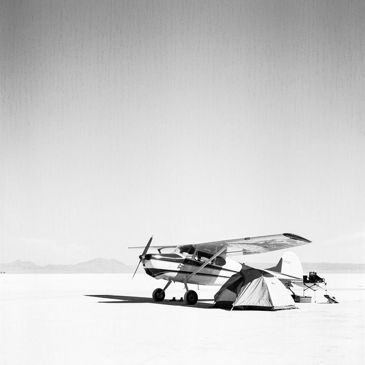 A small airplane is parked next to a tent on a vast, flat, and barren landscape with mountains in the distance.