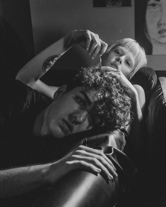 A man with curly hair rests his head on a woman's lap as she looks at the viewer.