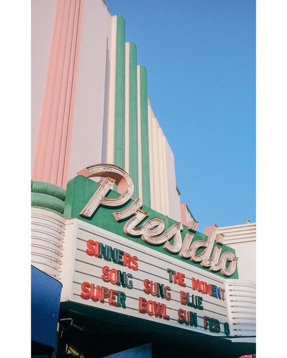 A movie theater marquee with the name "Presidio" is visible against a clear blue sky.