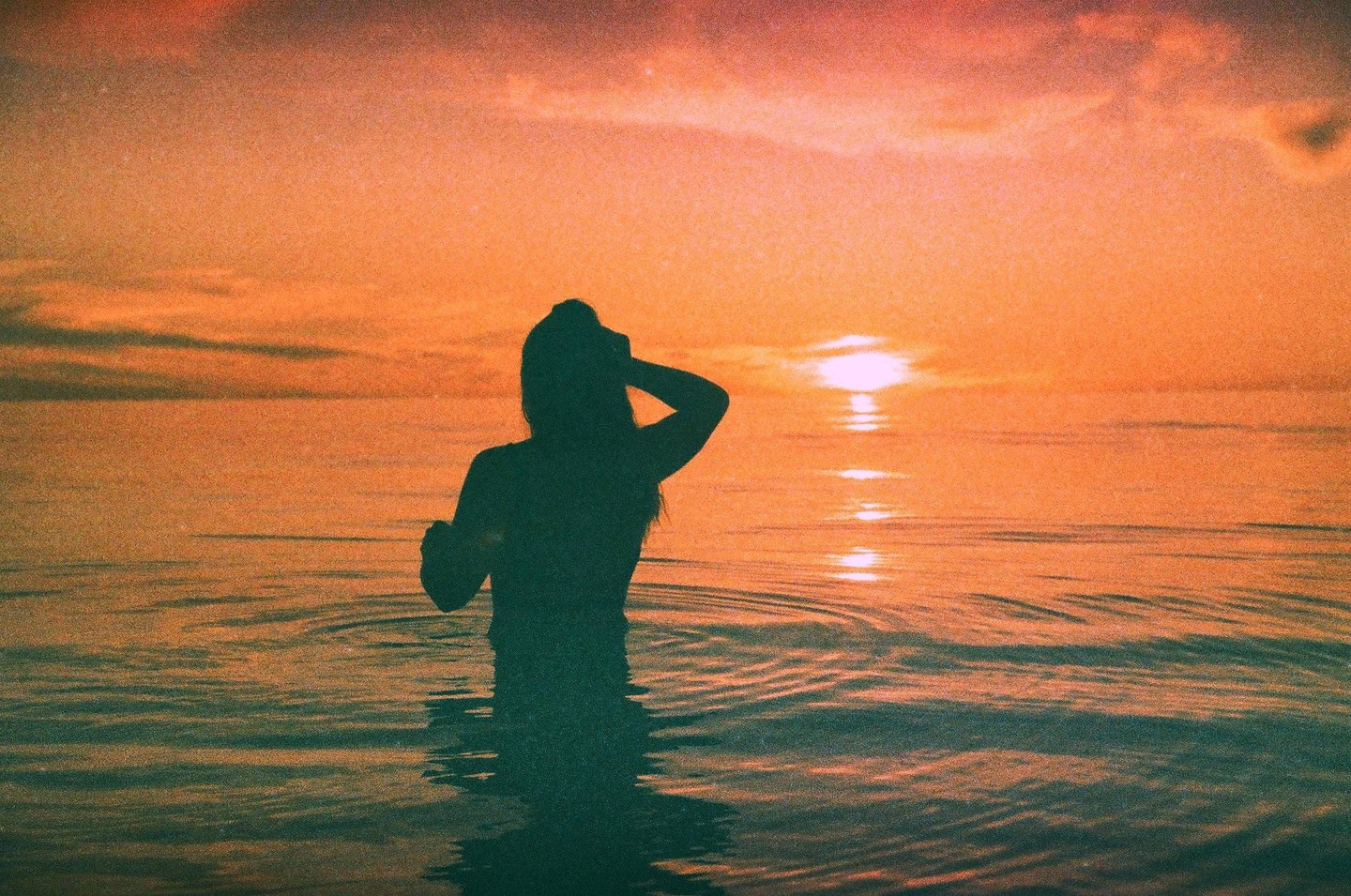 A woman stands in the ocean with the setting sun reflecting on the water behind her.