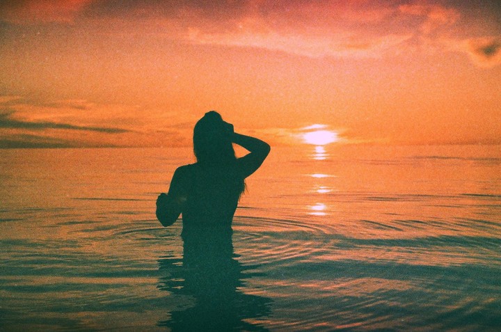 A woman stands in the ocean with the setting sun reflecting on the water behind her.