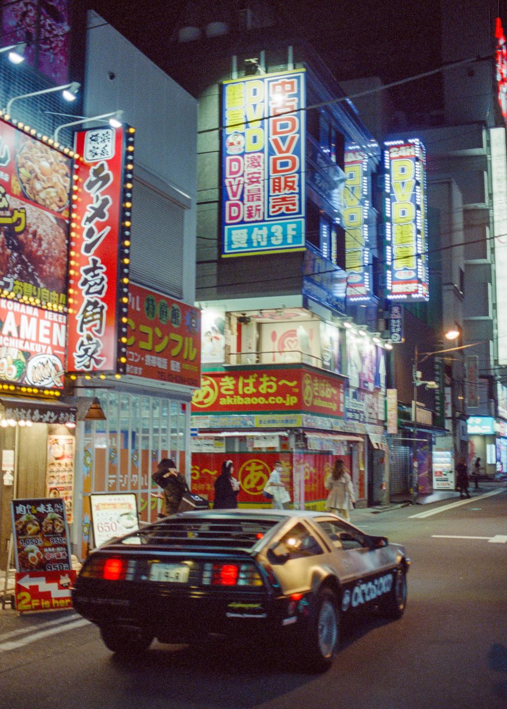 A DeLorean car is parked on a city street at night, surrounded by neon signs and buildings.