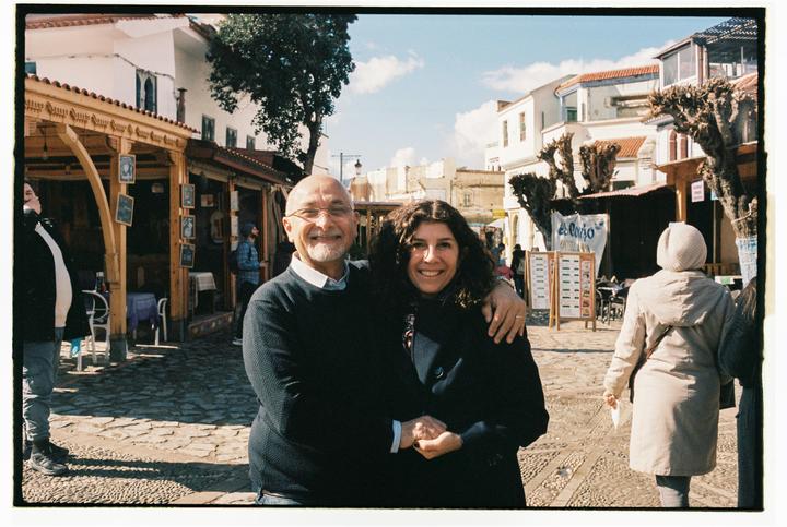 A man and a woman smile at the camera while standing on a cobblestone street lined with shops and buildings.