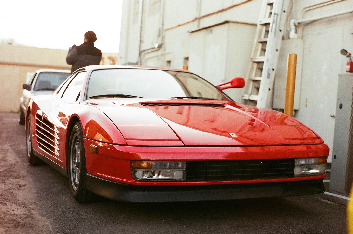 A red Ferrari Testarossa is parked on a street with a person standing behind it and a ladder leaning against a building in the background.