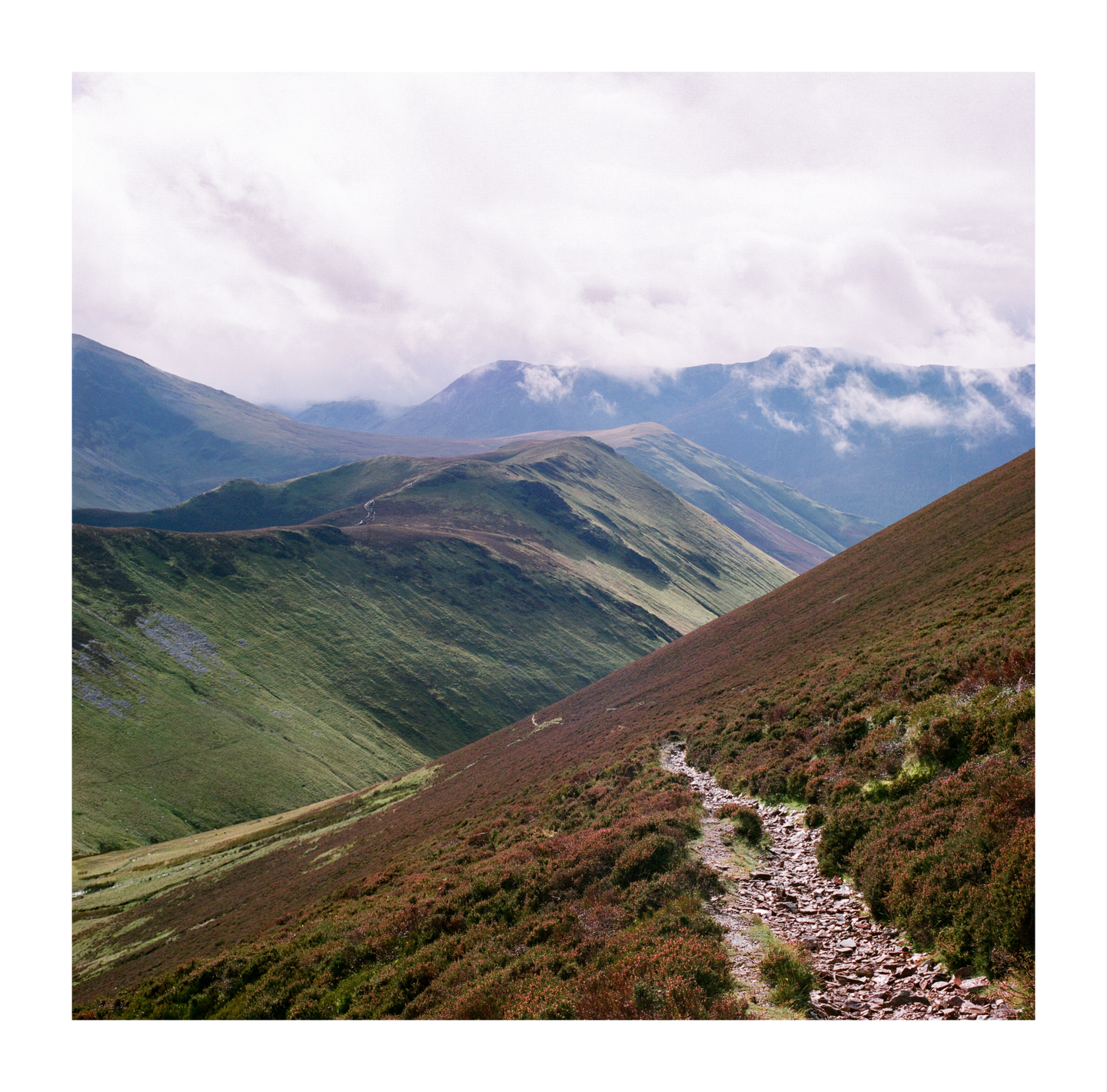 A rocky path winds up a steep, heather-covered hillside with green valleys and cloudy mountains in the background.