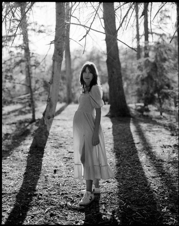 A woman stands in a forest with dappled sunlight and shadows on the ground.