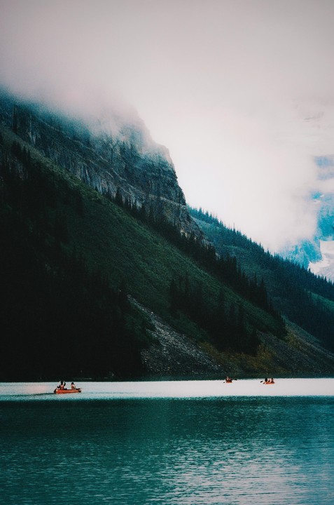 Three orange canoes with people are on a lake with a large mountain covered in fog in the background.