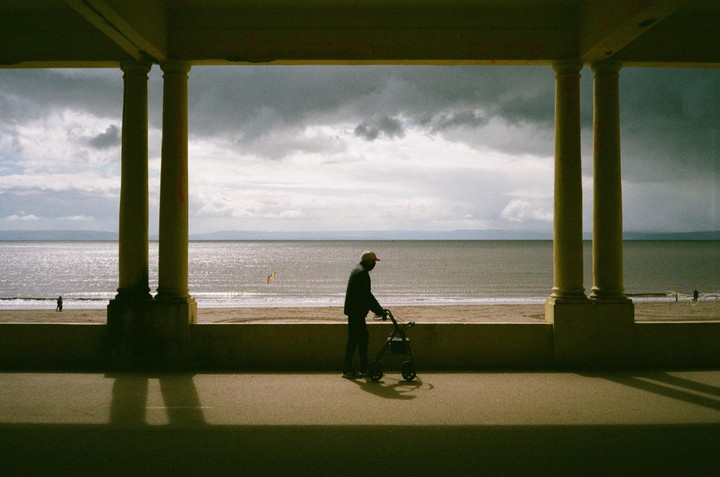 A man with a walker is silhouetted against a beach and ocean under a cloudy sky, framed by pillars.