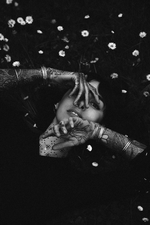 A woman with henna-decorated hands lies in a field of daisies, covering her face with her fingers.