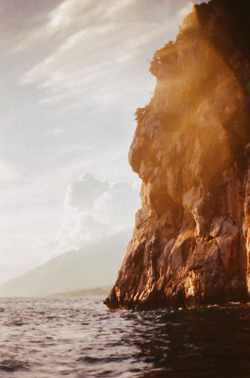 A sun-drenched cliff face rises from the ocean with mountains in the distance under a cloudy sky.