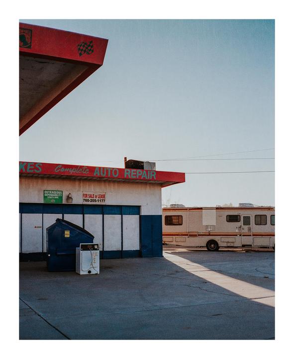 A recreational vehicle is parked next to an auto repair shop with a dumpster and washing machine outside.