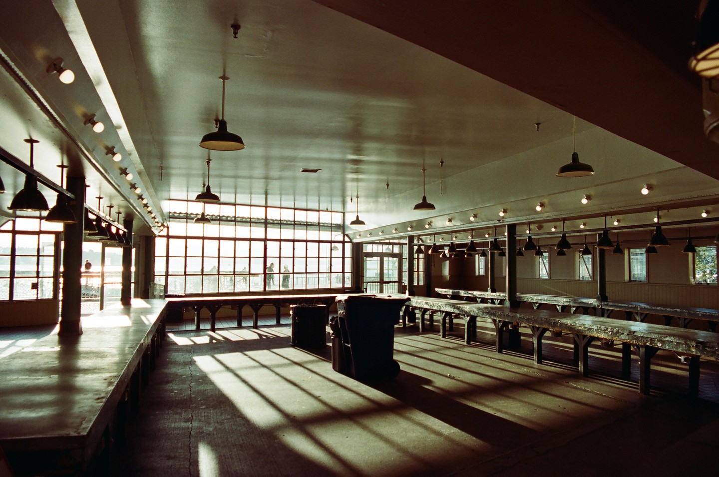 Sunlight streams through large windows into an empty market hall with long tables and hanging lamps.