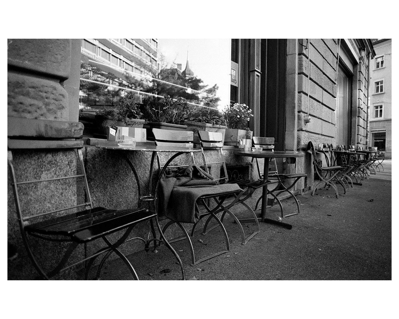 A row of empty cafe tables and chairs sits outside a building with reflections in the window.