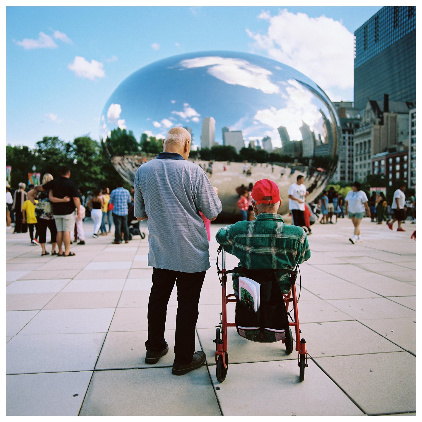 Two men stand in front of a large reflective bean-shaped sculpture in a city square.