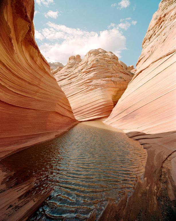 A narrow canyon with wavy sandstone walls is partially filled with water under a blue sky with clouds.