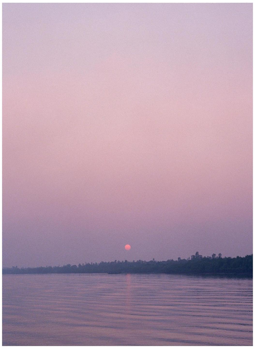 The sun sets over a body of water with a tree-lined shore.