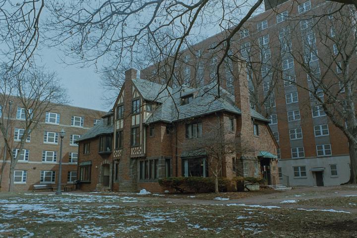 A brick building with a gray roof stands in front of a larger brick building, with bare trees and patches of snow in the foreground.