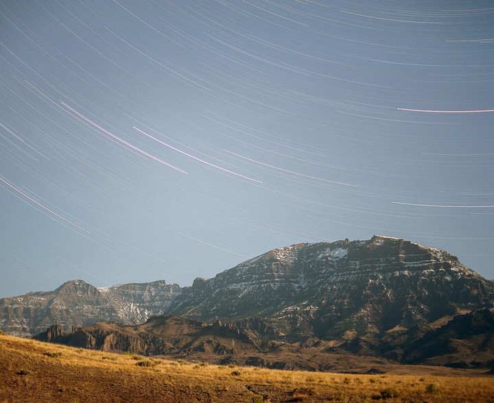 Star trails arc across the night sky above a mountain range with patches of snow and a grassy foreground.