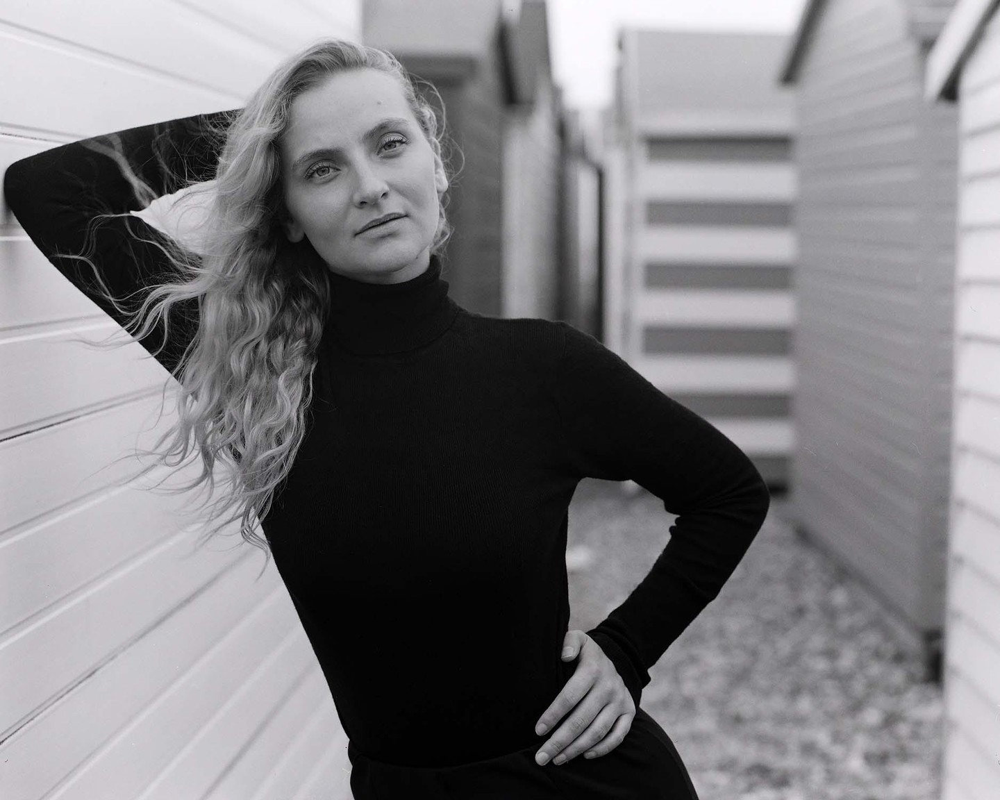 A woman with long, curly hair poses in a black turtleneck against a white wall with beach huts in the background.