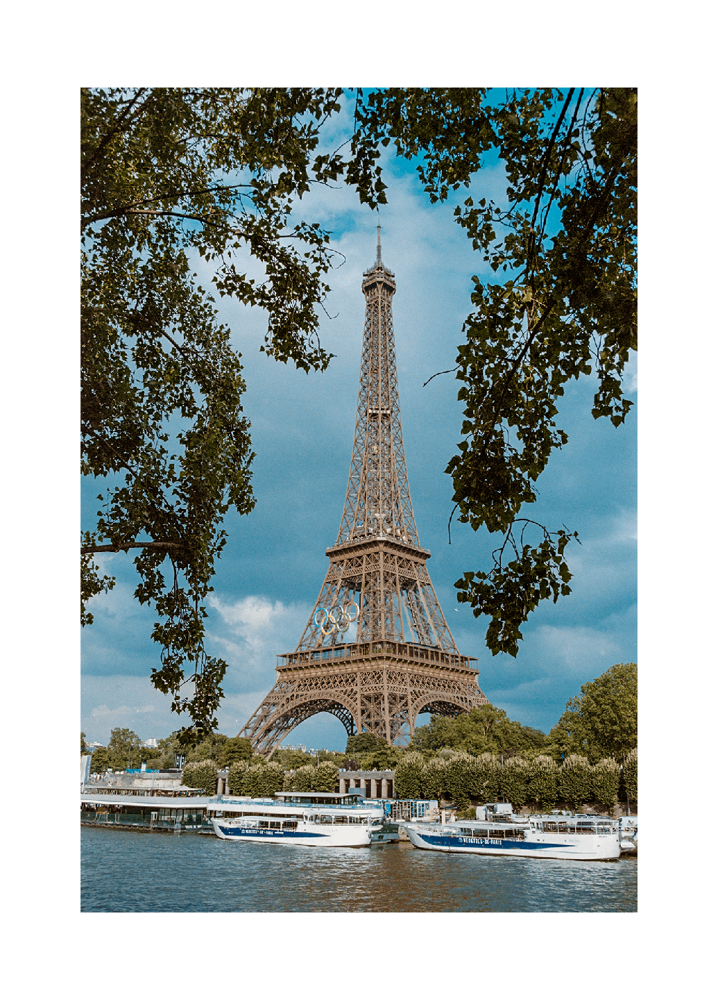 The Eiffel Tower is visible through tree branches, with boats docked on the river below.