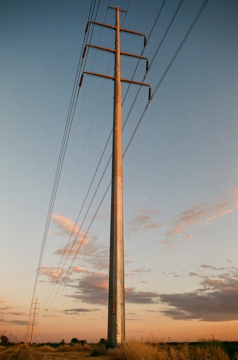 A tall power line pole stands in a field under a colorful sky at sunset.