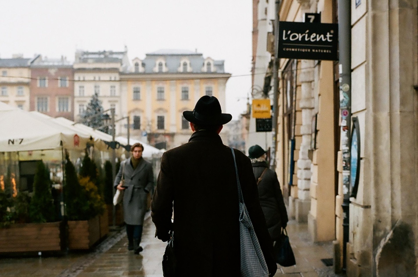 A man in a hat walks away from the camera on a city street with other pedestrians and buildings.