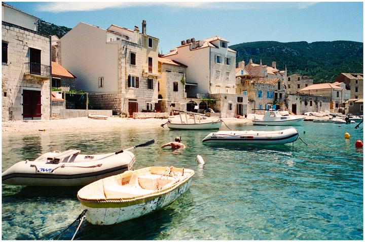 A man swims in clear blue water among boats near a village with stone buildings and a mountain in the background.