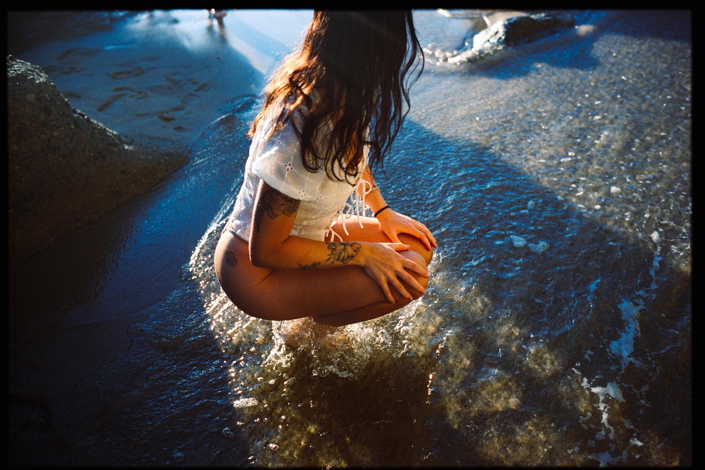 A woman crouches in shallow water at the beach, with sunlight reflecting on the water.